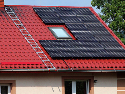 Tejado rojo de una casa con paneles solares instalados y una escalera apoyada, rodeado de árboles, representando energía solar y almacenamiento en batería virtual en Mallorca.